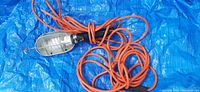 Photo of a portable trouble light with a protective metal cage and long coiled orange cord on blue tarp background.