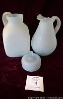 Photo showing two vintage frosted glass pitchers and one frosted glass dome lid on a burgundy cloth background, with a MaxSold lot number tag.