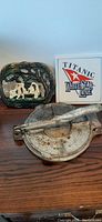 Photo of vintage tortilla press, Titanic ceramic trivet, and cast iron farm-themed trivet on wooden surface.