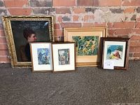 Five small framed art pieces lined up against a brick wall including a detailed portrait and colorful abstract prints.