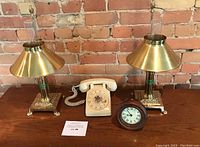 Shows pair of polished brass oil style lamps, vintage beige rotary telephone, and wooden mantle clock together on wooden table against brick wall.