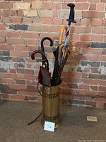 Full view of brass umbrella stand holding umbrellas, walking pole, and cane against a brick wall