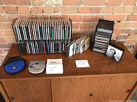 Photo showing about 90 CDs stored in two black racks, Sony Discman portable CD player with manual and carrying case on wooden surface with brick wall background.
