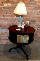 Wooden round accent table with a rotating top, styled as a lazy susan, with books in open side storage and a lamp and sculptures on top, against a brick wall.