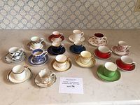 Overview of 16 assorted demi tasse cups and saucers arranged on countertop showing variety of colors and patterns.