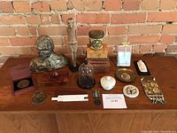Full lot of various decorative items including a bust sculpture, wooden boxes, figurines, brass and stone pieces, displayed on a wooden table against a brick wall.