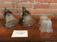 Pair of antique brass wall sconces with one included etched glass shade each and two additional glass shades displayed on wooden surface against brick wall.