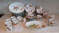 Full view of assorted Royal Worcester Evesham Vale patterned plates, cups, mugs, and serving platters with fruit motifs and green rims on a counter surface.
