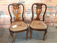 Set of two mahogany chairs with needlepoint upholstered seats featuring floral motif, shown against brick wall background.