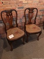 Photo of the pair of mahogany wood chairs with needlepoint floral upholstered seats against a brick wall.