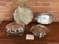 Full group shot of the 4 silver plated serving pieces against a brick wall background on wood surface.