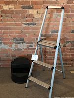Photo showing the three-step aluminum folding ladder alongside the black plastic library stool against a brick wall background.