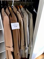 Five men's trench coats hanging on wooden and plastic hangers in a closet, mostly in tones of beige and tan.