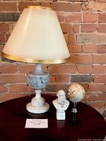 Full view of the lamp with marble base and gold-trimmed lampshade, bust sculpture, and shell sphere on table against brick wall
