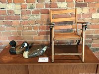 Wide shot of two wooden duck decoys displayed next to small oak folding child's chair on wood surface with brick background.