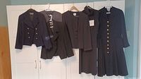 Full view of five vintage designer garments, four jackets and one dress, hanging on hangers against a white door.