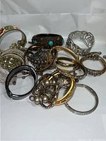 Twelve mixed metal bracelets in various styles and colors arranged together, showing details such as stones, crystals, and cutout designs.