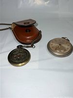 Two pocket watches placed on a white surface, showing the bronze watch with leather case and silver Westclox Pocket Ben watch side by side.