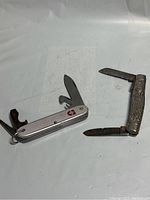 Two silver pocket cutting tools, one with textured tree-like handle featuring a crown emblem, the other with checkered silver handle and red lifeguard cross emblem, both with folding blades.
