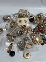 Top view of over 30 pairs of earrings sorted in small plastic bags showing a mix of gold, silver, and brass metals with varied styles including hoops, clip-ons, and costume pieces.