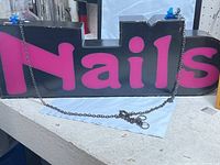 Front view showing 'Nails' spelled out in purple lettering on a black rectangular sign with silver hanging chain attached.