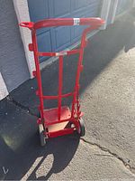 Red Smart Cart dolly standing upright on pavement in front of garage. Shows handle and rear wheels.