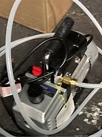 Vacuum pump with clear hoses and power cord visible from top and side angles, showing red valve knob and black circular cap.