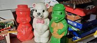 Front view showing three colorful 12-inch blow mold character banks - red Porky Pig, white bear, and green Ninja Turtle, arranged side by side on a surface with background boxes and items.