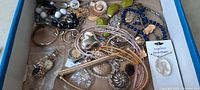 Photo showing the entire jewelry lot inside a box with assorted jewelry pieces including necklaces, bracelets, brooches, rings, and a wristwatch.
