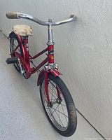 Front left side view of red antique kids bicycle showing chrome handlebars, worn cream seat, front wheel and fender.