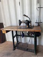 Front angle view of vintage Singer treadle sewing machine on wooden table with foot pedal and lamp attached.