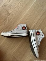Side view of two Converse high-top sneakers showing gray canvas with orange interior and black Converse All Star logo patch. White rubber toe caps and midsoles. Shoes are missing laces.