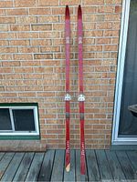 Full view of both vintage wooden cross country skis showing red color, metal bindings, and overall length.
