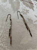 Pair of dangling earrings resting on a light fabric background. The earrings have thin chain strands with small bar details hanging vertically. Fishhook ear wires attached.