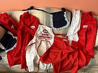 Photo showing three red long coats with Longo's patches, two navy blue aprons with frilly edges, the white IGA Beef Roundup apron, and white scarf, laid out on a sofa