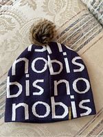 Front and top view of Nobis branded dark blue and white knit toque with faux fur pom pom on top, displayed on beige patterned fabric surface.