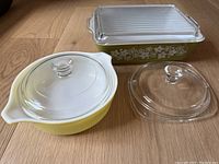 Overall lot showing rectangular dish with floral design and ribbed glass lid, round yellow casserole with dome lid, and extra glass lid