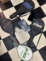 Four Prince tennis racquets with three black soft cases and one green-white hard case, set of golf tees, Wilson golf balls and black golf club cozy on black and white tiled floor