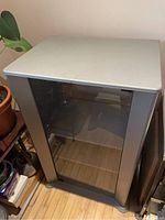 Front view of the stereo cabinet showing the gray top with the transparent front door closed, revealing empty compartment inside.