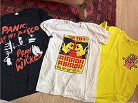 Three t-shirts laid flat on floor: black Panic! At the Disco, white Star Trek, and yellow Keith Haring inspired tee