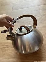 Side angle showing the kettle with curved wood handle, metal spout with whistle, and wood lid knob on wood floor.