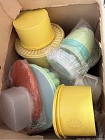 Box full of various vintage Tupperware containers and jello molds, mostly yellow, green, orange, and opaque plastic, some wrapped in plastic.