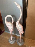 Full view of two vintage bird statues standing side by side on wooden floor against grey background