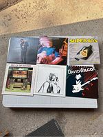 Six vinyl albums laid out on a plastic container in natural light with various cover arts including a portrait of a man with a guitar, an abstract motion blur image, and a colorful 60s themed design.