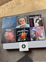 Seven vinyl record album covers and a small CD, arranged on a white plastic table outdoors showing diverse artists and genres.