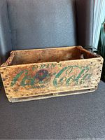 Front side view of the wooden Coca-Cola crate showing faded green "Drink Coca-Cola" lettering and wooden texture with visible wear and cracks.