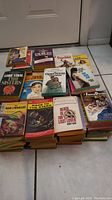 Photo shows stacks of paperback novels on tiled floor against white door. Titles include 'The Exorcist,' 'Anne Frank,' 'Texas Rebel,' and more, indicating a large collection of adult themed novels with some covers showing age and wear.