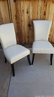Pair of off white/off eggshell leather dining chairs with slightly curved backs and dark wooden legs against wood panel background.