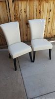 Two white leather dining chairs with black legs, placed side by side on concrete with a wooden fence background.