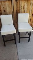 Pair of two off white/eggshell leather barstools with dark wood legs and footrests against wooden fence background.